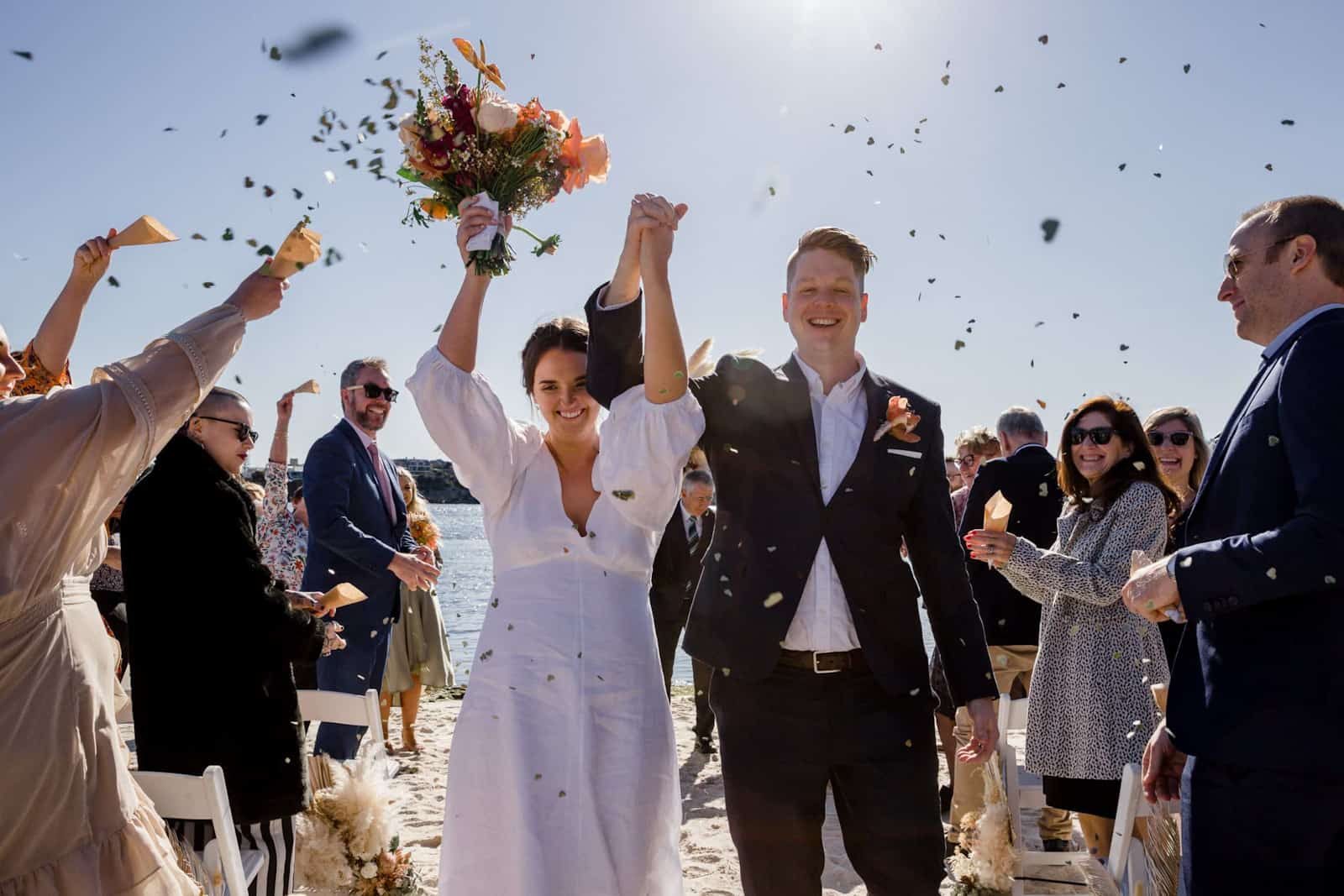 newly married couple celebrate with hands in the air as they walk down the aisle as guests throw confetti